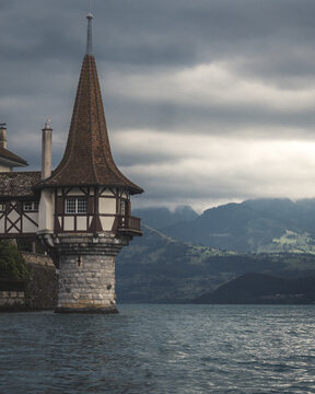 View of a medieval castle's stone tower adorned with a pointed roof, reflected in the still waters of a lake under a brooding sky, Oberhofen am Thunersee, Bern, Switzerland.