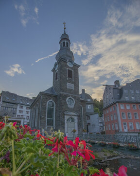View of the historic Monschau Protestant Church rises majestically, framed by vibrant red geraniums and the serene Rur river, Monschau, North Rhine-Westphalia, Germany.