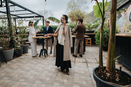 A diverse group of professionals socializes on a rooftop patio, talking on phones and standing by high tables among potted plants. The casual business atmosphere blends urban scenery with greenery. - Powered by Adobe