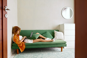 Young woman working on a laptop at home, relaxing on a green sofa in a cozy room, showcasing a modern lifestyle with a casual outfit and a peaceful atmosphere