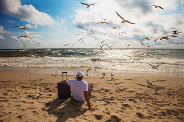 A human sits on the beach in summer with a travel suitcase. The man looks at the sea. Seagulls fly...