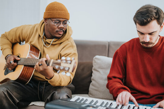 Two friends collaborate musically, one strumming a guitar and the other playing a keyboard. They sit on a sofa in a bright, casual home setting.