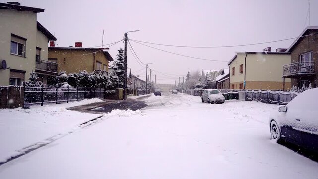 Snowy residential street during snowfall with detached houses parked cars limited visibility in quiet suburban neighborhood area