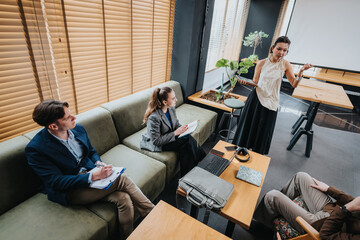 A group of colleagues participates in a business meeting in a contemporary office. One presenter stands while others take notes and engage, with laptops, notebooks, and plants nearby.