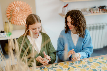 Two friends sit together in a cozy, artistic studio, shaping clay into unique forms. Surrounded by creative tools, they share laughter and inspiration while working on their pottery.