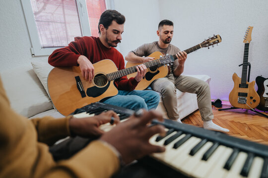 Three friends play acoustic guitars in a cozy living room. A keyboard player in the foreground provides rhythm as another guitarist sits on a couch beside a wall of guitars.