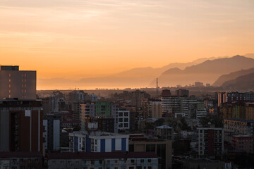 Fototapeta premium Urban cityscape at sunset with warm golden sky, modern residential buildings, layered mountains in background, evening atmosphere, calm mood, travel and real estate concept