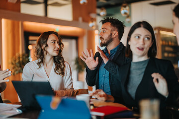 A diverse group of colleagues gathers around a desk, engaging in an animated discussion with laptops and notebooks in a stylish, well-lit office.