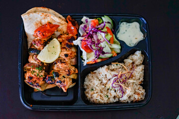 Top view of grilled chicken with pita bread, rice, fresh vegetable salad and yogurt sauce in a takeaway meal box. Healthy Mediterranean or Middle Eastern food concept.