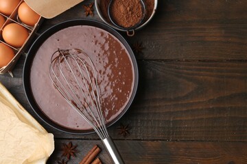 Liquid chocolate dough in bowl, whisk and ingredients on wooden table, flat lay. Space for text