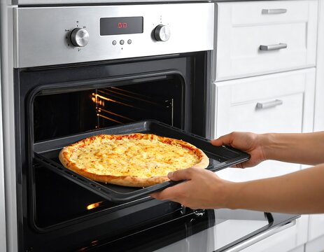 Person sliding a golden, cheesy pizza on a dark pan into an open oven in a bright white kitchen setting