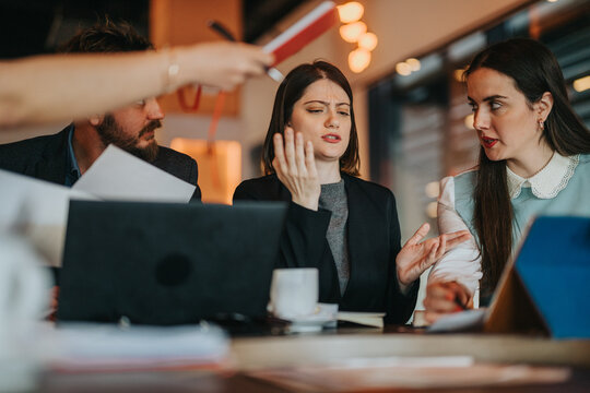 A tense meeting among colleagues around a cluttered table with laptops and papers; distressed gestures and focused faces convey disagreement and collaboration in a modern work environment.