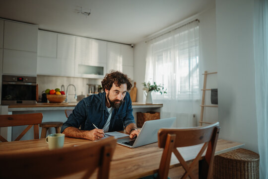 Man working remotely using laptop and notepad at home kitchen table