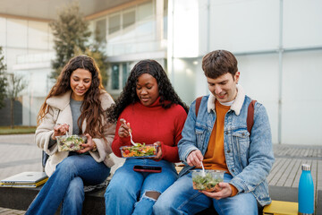 Diverse young people sitting outdoors, eating fresh salads and sharing a meal