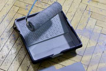 Close-up showcasing a paint roller soaked in gray paint, resting on a paint tray placed on a wooden parquet floor splattered with gray paint, highlighting a home improvement task.