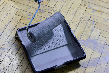 A paint roller and tray filled with gray paint rest on a herringbone wooden floor, prepared for painting. This image represents interior house renovation, do-it-yourself initiatives, and the