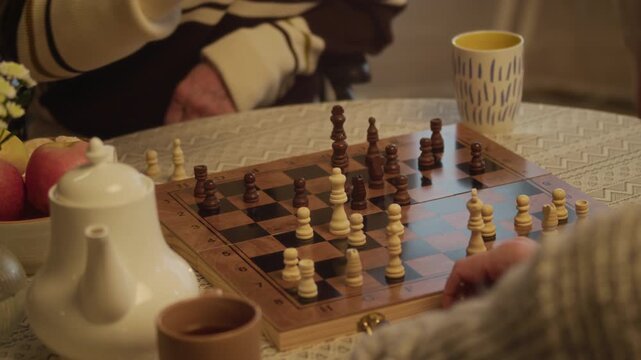 Tilt up shot of elderly Caucasian man sitting at table with teapot and mugs, playing chess with unrecognizable companion focused on next move in cozy nursing home setting