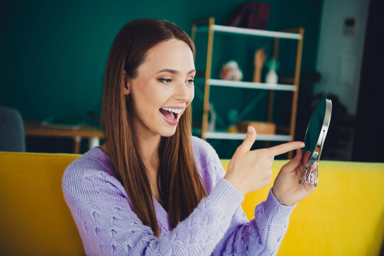 Happy brunette woman in a cozy purple sweater smiles while showing a mirror in a bright modern living room at home with yellow couch - Powered by Adobe