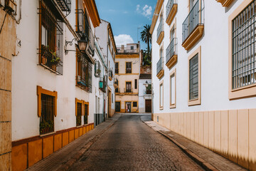 Traditional southern Spanish architecture lining a narrow cobblestone street. Highlighting white washed buildings with yellow trim and wrought iron balconies under a clear blue sky in historic Cordoba