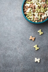 A blue bowl filled with colorful bone shaped dog treats on a gray background, with some scattered outside.