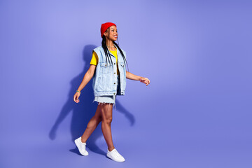 Young woman walks confidently in denim vest yellow tee red beanie white sneakers against violet...