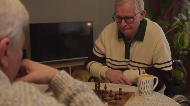 Over-the-shoulder slowmo shot of grey-haired elderly Caucasian male friends in cozy sweaters focused on playing chess match spending evening together in home setting