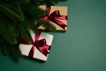On a green background, under spruce branches, lie two gift boxes tied with red satin ribbons