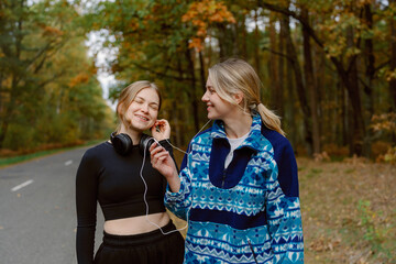 Fototapeta premium Two friends enjoy a joyful moment while walking along a peaceful road in the forest. Wearing cozy outfits, they share headphones, smiling and immersed in their music as autumn leaves surround them.