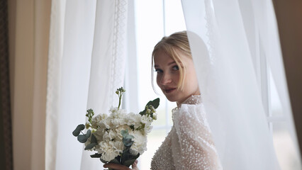 Beautiful bride holding a white bouquet and looking over her shoulder while standing near a window...