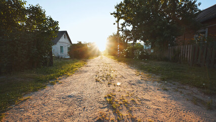 Sunlight streaming over a dirt road at sunset, illuminating a peaceful countryside scene with a wooden fence and charming houses