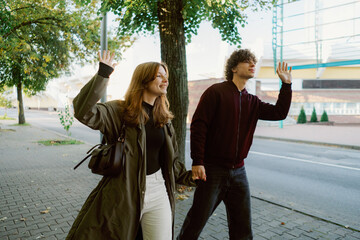 A young couple walks together under a clear sky, waving happily at passersby. The trees provide gentle shade while they enjoy a peaceful moment in the heart of the city.