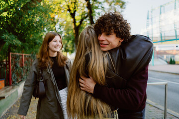 Obraz premium Two friends joyfully hug outside, surrounded by trees showing autumn colors. A third friend smiles nearby, capturing the moment in a vibrant urban setting.