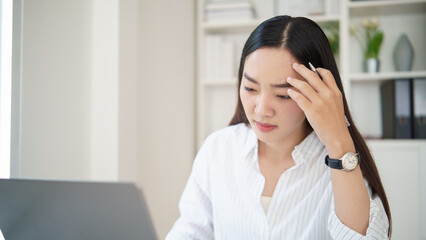 Fototapeta premium Concerned business experiencing stress at work, touching her forehead while looking at a laptop in a bright home office.