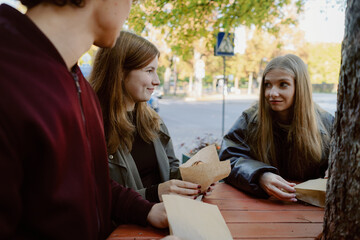 Three friends sit at a wooden table under trees, exchanging smiles while enjoying snacks. The sun shines warmly, creating a relaxed atmosphere in the park.