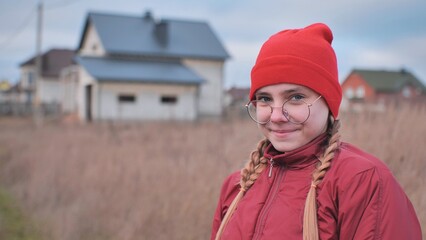 Teenage girl smiling, wearing red beanie and glasses, standing in grassy field with residential neighborhood behind