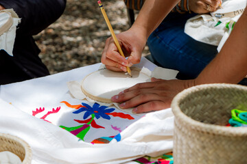Close-up of hands designing a pattern with a pencil on embroidery fabric with a nearby traditional Mexican floral design.