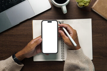 Top view of hands holding smartphone with blank white screen over open spiral notebook, perfect mockup for app or branding.