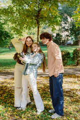 Fototapeta premium Three friends pose cheerfully in a vibrant park filled with green trees and fallen leaves. They laugh and capture memories together, enjoying the warm sunlight of a beautiful afternoon.