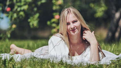 Blonde woman resting on soft blanket amid fruit trees, basking in golden sunlight during peaceful summer afternoon in verdant orchard