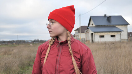 Young girl sporting braided hair, red beanie, winter jacket, gazing across rural landscape with unfinished residential construction site behind her