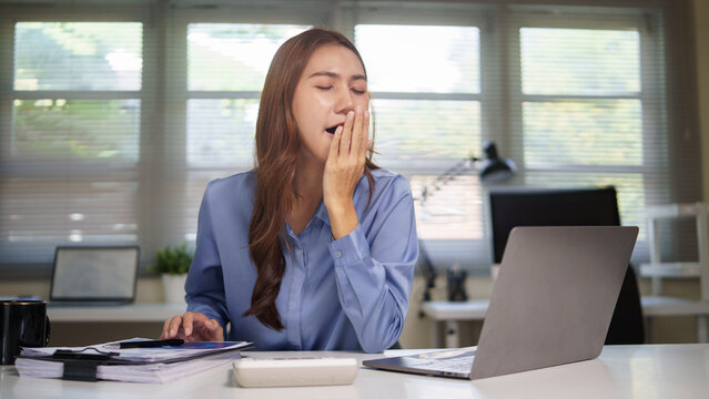 Young businesswoman feeling sleepy at work, yawning at her desk in a modern office.