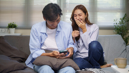 Happy young couple sitting on a sofa at home, laughing together while watching something on a...