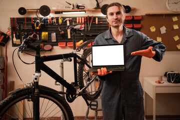 Male bicycle mechanic pointing with pointer finger on laptop computer with blank screen workspace area while working in his repair workshop