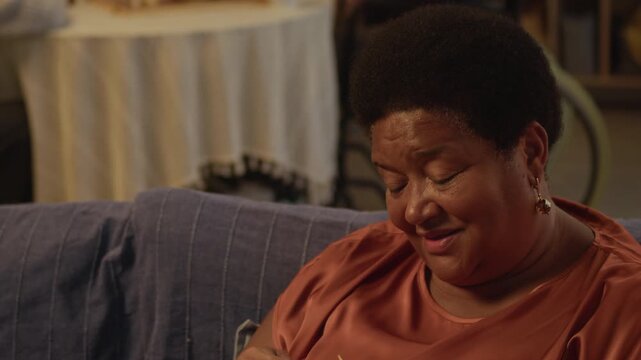 Rack focus slowmo shot of smiling elderly African American woman knitting on couch spending peaceful evening in cozy nursing home with Caucasian male seniors playing chess in background