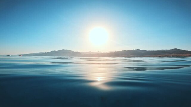 Calm hypnotic sea water surface with sunlight reflection and distant coast with mountains under clear sky during sunset. Low angle cinematic shot
