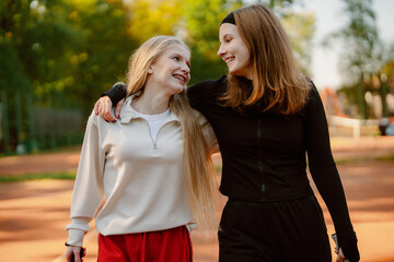 Fototapeta premium Two friends walk side by side in the park, smiling and chatting as they enjoy the sunny weather. Their laughter fills the air as they share a joyful moment after exercising.