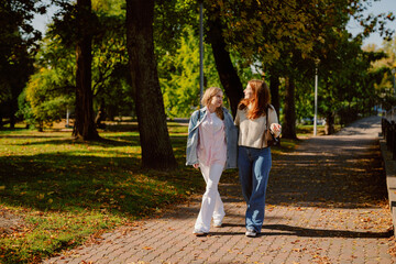 Fototapeta premium Two friends stroll through a beautiful park on a sunny day, surrounded by lush greenery and falling leaves. They share smiles and laughter, creating joyful memories together.