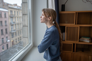 Dreaming woman enjoys looking at window, mindful pause at home deep in pleased thoughts. Happy young female taking break spending slow time thinking about holidays. Calmness, gratitude, contentment.