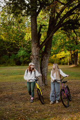 Fototapeta premium Two friends chat while walking their bikes through a park on a bright autumn day. The vibrant colors of the trees create a warm and inviting atmosphere around them.