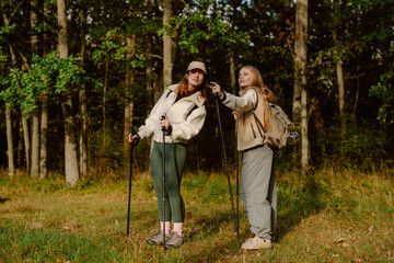 Obraz premium Two young women enjoy a lively hike through a forested area. They discuss the scenery while using trekking poles, surrounded by lush greenery on a beautiful day.